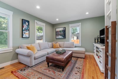 A stylish living room with a gray sectional, leather ottoman, paintings, and natural light.