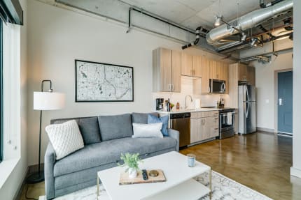 A modern apartment with a living area and kitchen, showcasing a gray sofa, floor lamp, and stainless steel appliances.