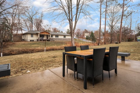 A modern outdoor dining set on a concrete patio overlooking a yard with a house and trees in the background.
