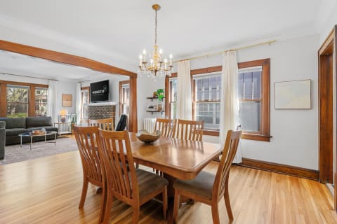 A dining table with wooden chairs and a chandelier, connected to a cozy living area with a couch and a fireplace.