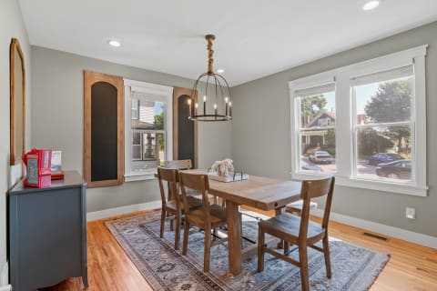 A cozy dining room featuring a wooden table, chairs, a chandelier, and large windows.
