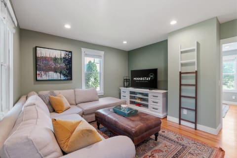 A cozy living room featuring a sectional sofa, a leather ottoman, a TV, and a lively painting.
