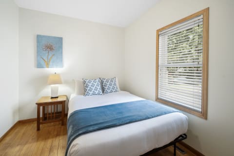 A cozy bedroom with a bed, lamp, and window, featuring soothing colors and decor.