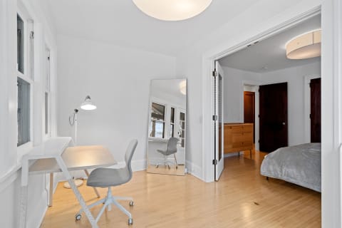 A bright, minimalist workspace with a light wood desk, grey chair, and mirror, leading into a bedroom area.