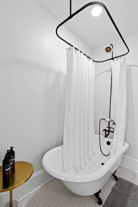 A vintage bathtub with white curtains and a golden side table holding bath products in a modern bathroom.