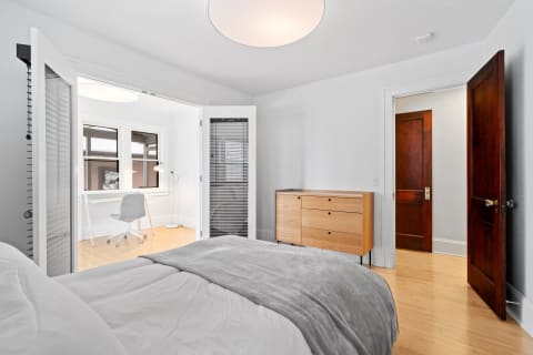 A minimalist bedroom featuring a bed, a wooden dresser, and a workspace with a desk and chair.
