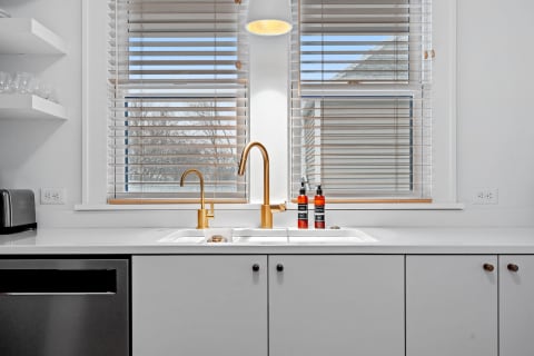 A minimalist kitchen countertop with a white sink, gold faucet, and amber bottles, illuminated by sunlight.