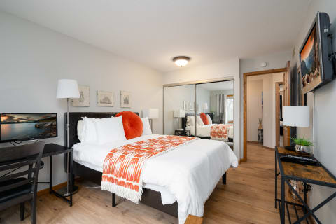 A bedroom with a white bed, orange throw blanket, and work area with a computer.