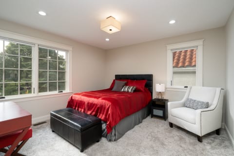 A well-decorated bedroom featuring a red bed, a nightstand, and a comfy chair by the window.