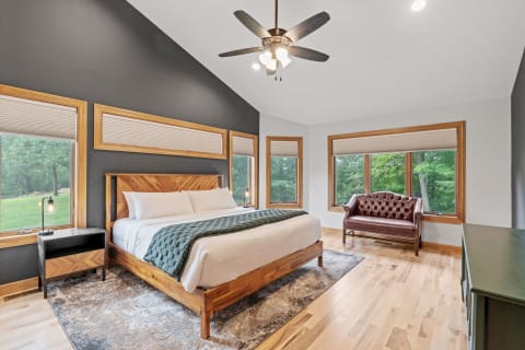 Modern bedroom featuring a wooden bed, leather loveseat, and large windows with views of greenery.