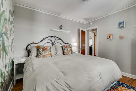 A serene bedroom featuring a large bed with gray bedding, decorative floral pillows, a white nightstand with a lamp, and a tropical mural on the wall.