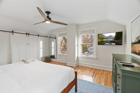 A serene bedroom with hardwood floors, a white bed, a green dresser, and a view of nature through the window.