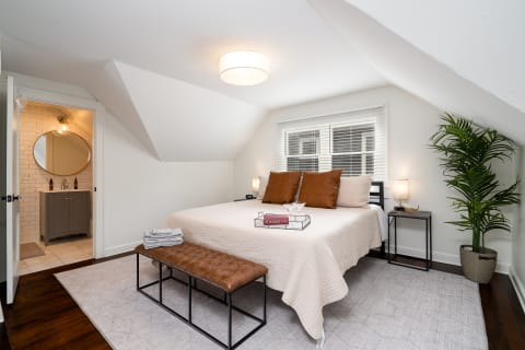 A spacious bedroom featuring a queen-size bed, a leather bench, and a small bathroom doorway in the background.