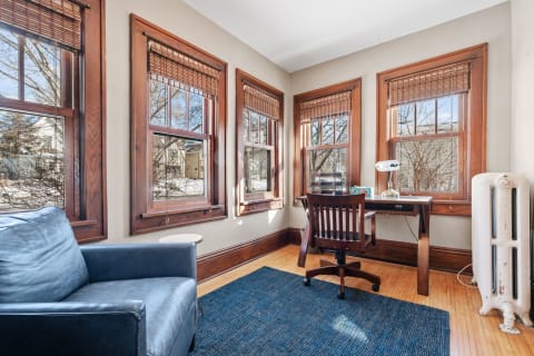 A bright home office with large windows, wooden furniture, and a blue rug.