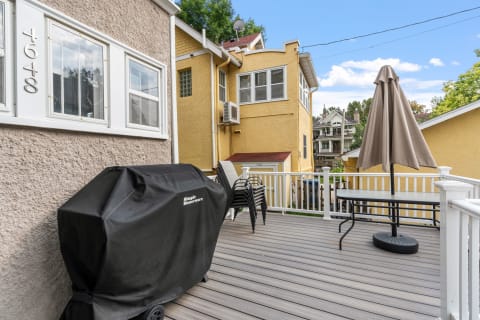 A deck with a grill, stacked chairs, and a table with an umbrella, set against yellow houses.