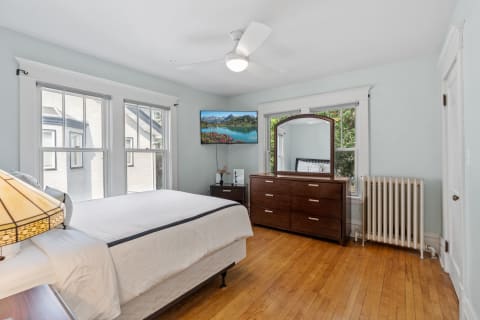 A cozy bedroom featuring a bed, dresser, large windows, and a mounted TV.