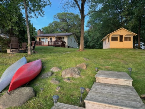 Lakeside view with canoes, a yellow cabin, and a larger house on a grassy hillside.