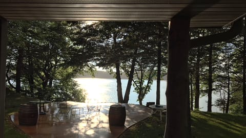 Lakeside patio with wooden barrels and metal chairs surrounded by trees.