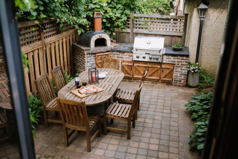 An outdoor patio with a wooden dining table, pizza, wine, and a barbecue grill in a lush garden setting.