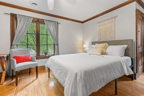 A serene bedroom with a neatly made white bed, gray curtains, and a cozy armchair.
