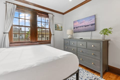 A bright bedroom featuring a bed, a dresser with a lamp, a TV showing boats, and large windows with curtains.