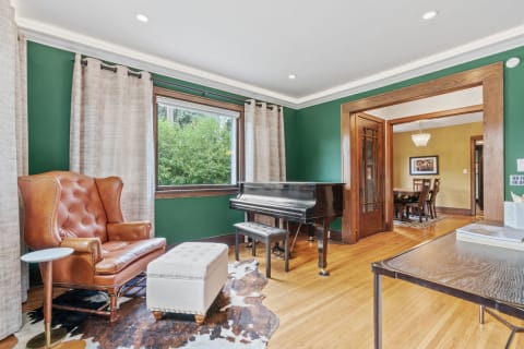 An interior room featuring a piano, leather armchair, and green walls, with bright natural light coming through the window.