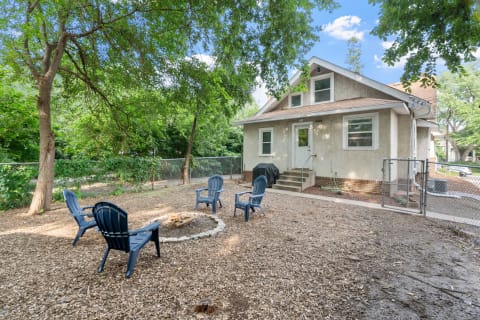 A backyard featuring a house, fire pit, and Adirondack chairs among greenery.