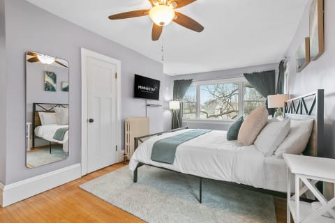 A cozy bedroom featuring soft lavender walls, a modern ceiling fan, and large windows with dark green curtains.