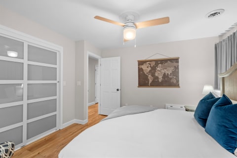 A modern bedroom featuring a queen-sized bed with blue pillows, a world map on the wall, and a ceiling fan.