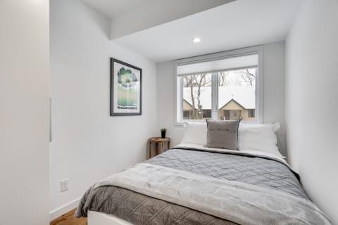 Minimalist bedroom with a grey bedspread, white pillows, and a large window overlooking snowy houses.