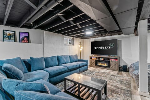 A cozy basement living room with a blue sectional sofa, artwork on the walls, and a television displaying a logo.