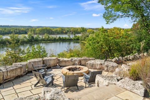 Outdoor fire pit with chairs, overlooking a river and forest.