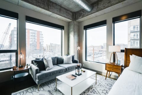 Interior of a modern living room featuring a gray sofa, white coffee table, and large windows with urban views.