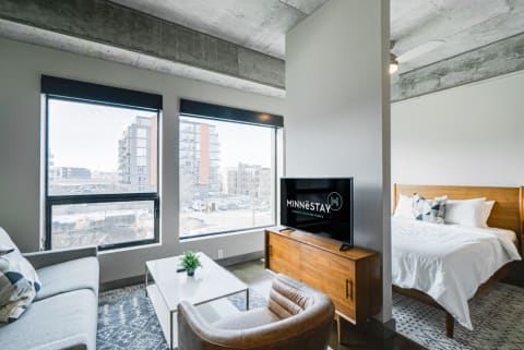 Interior of a modern studio apartment with a living area, bed space, and large windows showing an urban view.