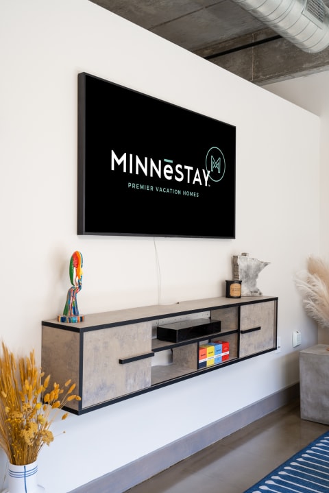 A stylish living room with a wall-mounted TV displaying the MINNĒSTAY logo, a sleek console table, colorful decor, and dried flowers.