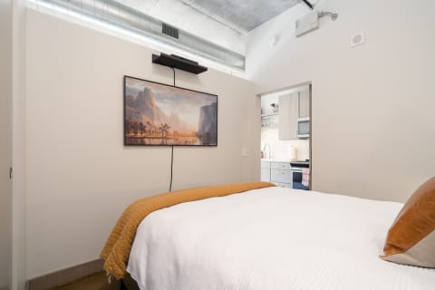 Comfortable bedroom featuring a white bedspread and mustard throw, a mounted TV, and a glimpse of a kitchen.