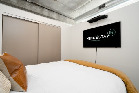 A modern bedroom featuring a bed with a white comforter and a golden throw, with a mounted TV displaying MINNESTAY logo.
