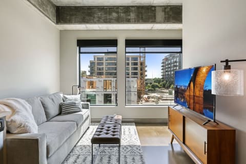 A contemporary living room featuring a gray sofa, an ottoman, and a large TV, with urban construction visible outside.