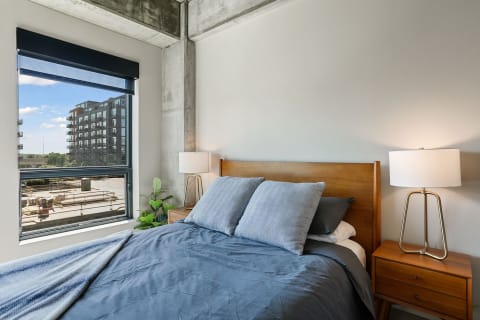 Contemporary bedroom featuring a large window, blue bedspread, and wooden nightstand.