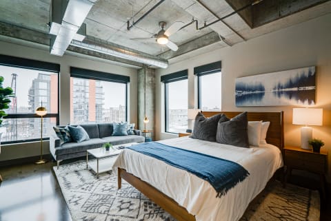 A modern urban bedroom featuring a bed with white linens, a gray sofa, and large windows overlooking a cityscape.