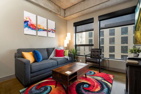 Modern living room with a gray couch, colorful pillows, abstract art, and large windows.