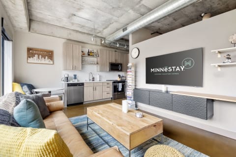 Contemporary living room and kitchen area with a cozy sofa and vacation rental logo on TV.