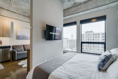 Interior of a contemporary studio apartment featuring a bed, sofa, kitchenette, and large windows.