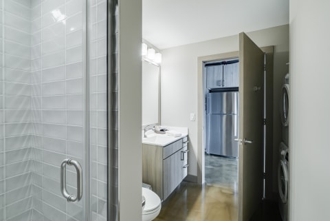 Interior view of a modern bathroom with a glass shower, vanity, and a glimpse of a kitchen.