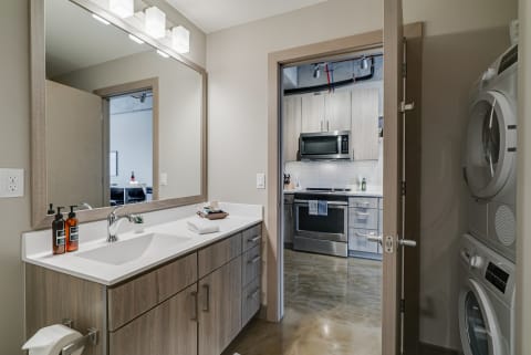 Interior view of a modern bathroom with a mirror, sink, and an adjoining kitchen visible.