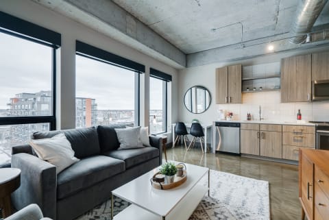 Interior of a modern apartment featuring a gray sofa, window views, and a kitchen area.