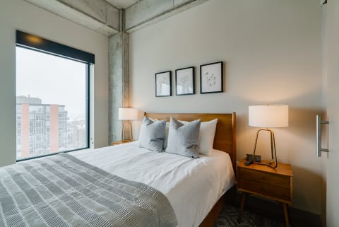 A well-decorated bedroom featuring a queen-sized bed, artwork, and a large window with city views.