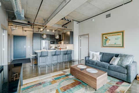 A modern living room with a kitchen area, gray sofa, and colorful area rug.