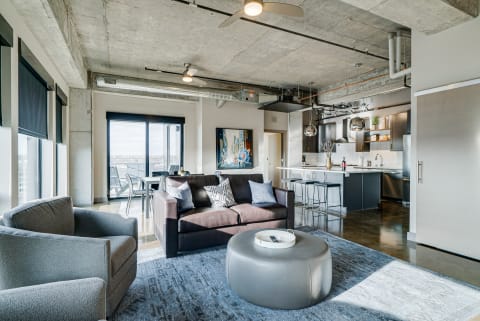 Contemporary living room and kitchen in a stylish apartment featuring a dark sofa, gray chair, and large windows.