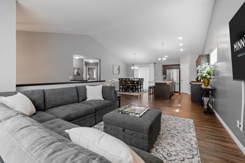 A modern living room with a gray sectional couch, an ottoman, and a dining area in the background.